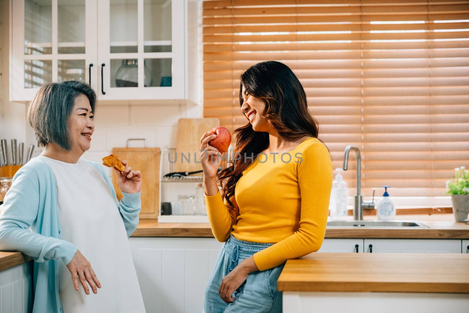 A cheerful Asian woman, with her mother, stands in the kitchen, holding an apple. Their warm embrace embodies the joy of teaching and learning within the family.