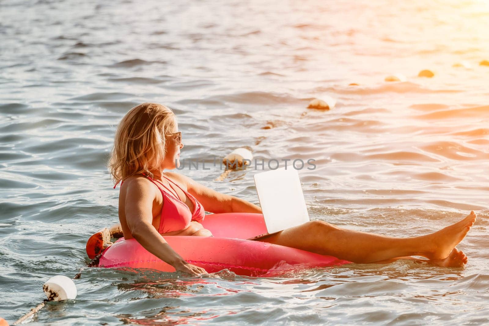 Woman works on laptop in sea. Freelancer, young blond woman in sunglases floating on an inflatable big pink donut with a laptop in the sea at sunset. Freelance, travel and holidays concept