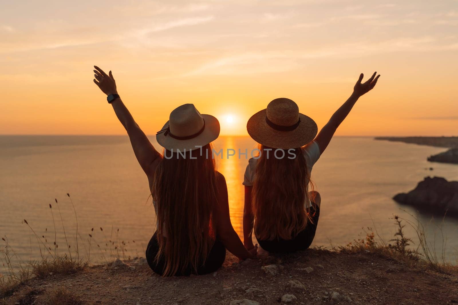 Mother daughter sunset. Back view of mother and daughter looking at sunset. Couple sitting back on a beach with their hands raised, watching and enjoy the sunset. by Matiunina
