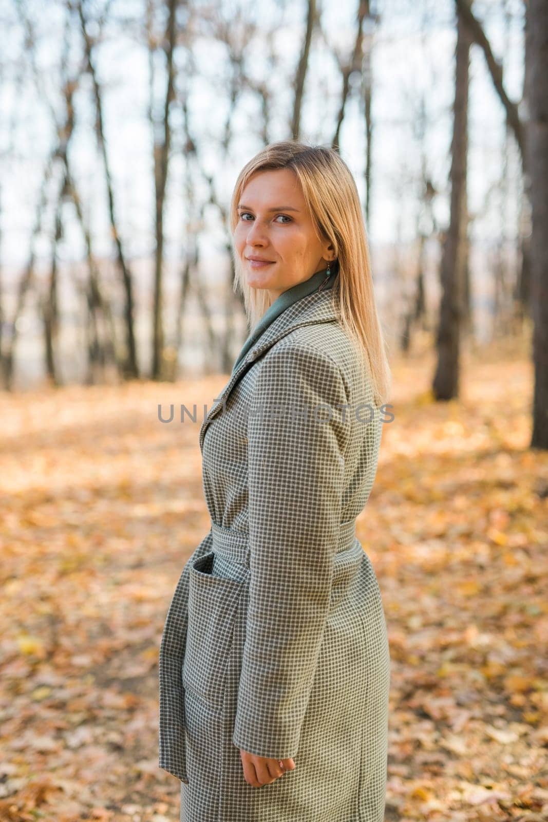 Blonde woman in elegant gray coat walks in the sunny autumn season park. Generation z and gen z youth concept