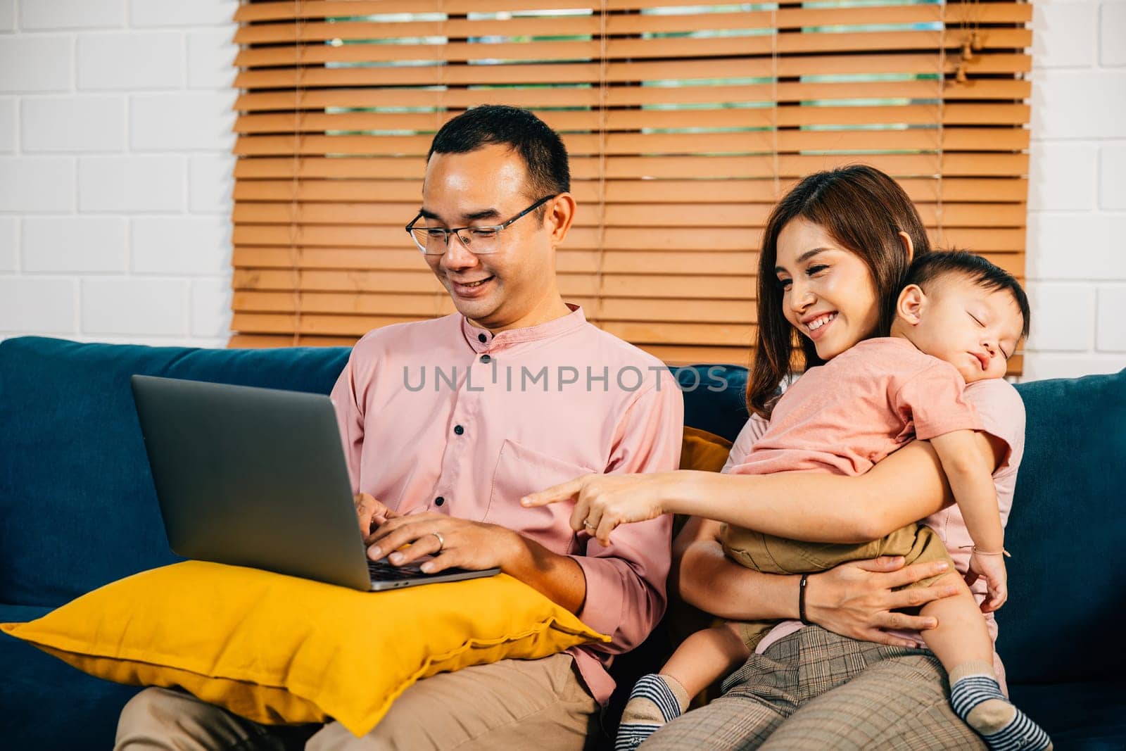 A businesswoman carries her sleeping daughter while her husband works on a laptop by Sorapop