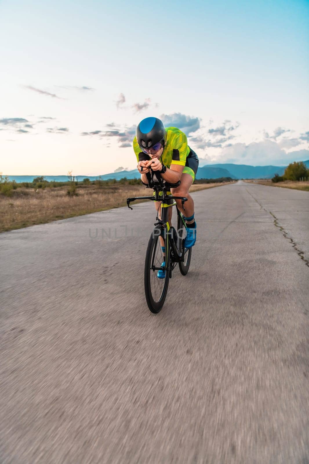 Triathlete riding his bicycle during sunset, preparing for a marathon. The warm colors of the sky provide a beautiful backdrop for his determined and focused effort. by dotshock