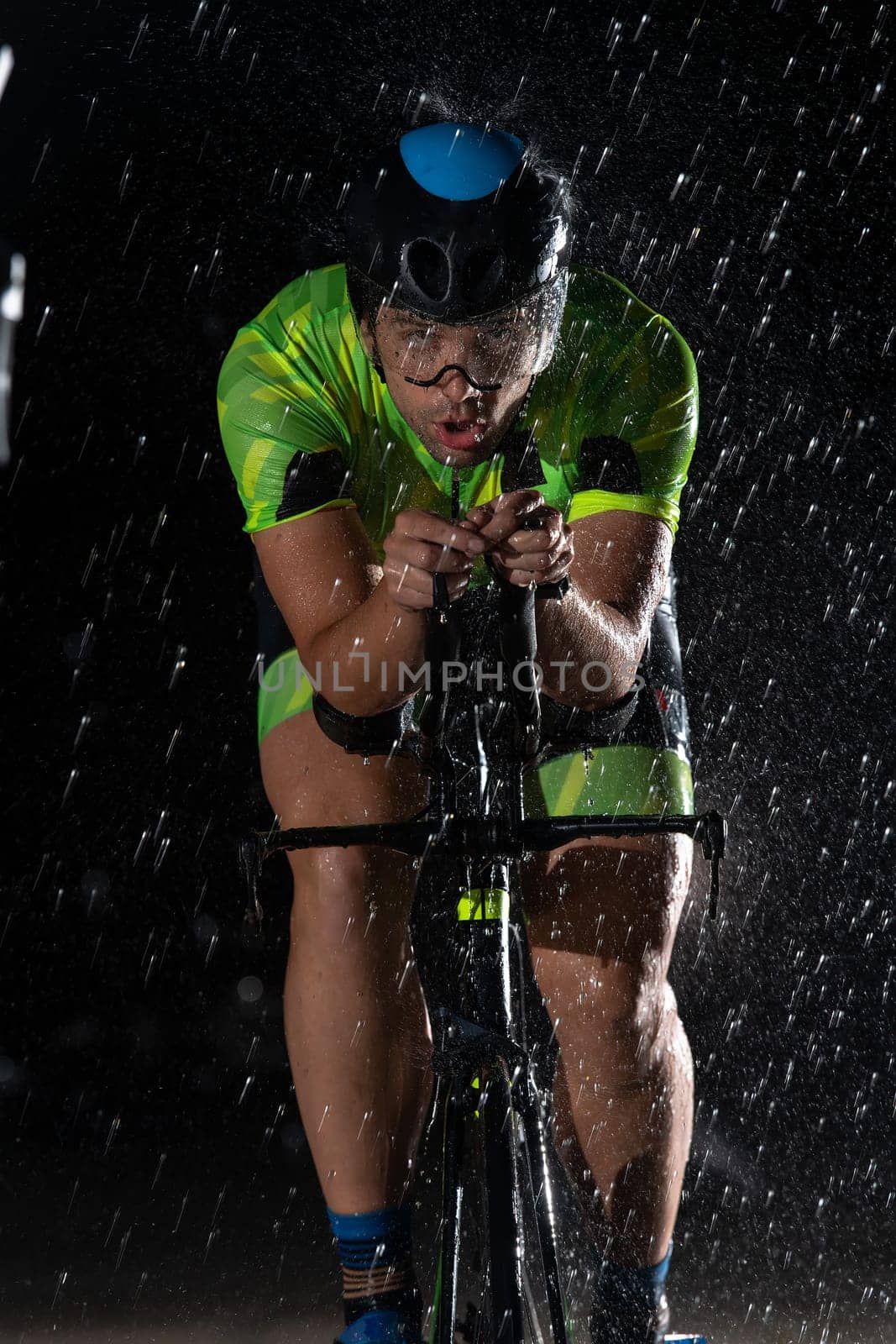 A triathlete braving the rain as he cycles through the night, preparing himself for the upcoming marathon. The blurred raindrops in the foreground and the dark, moody atmosphere in the background add to the sense of determination and grit shown by the athlete. by dotshock