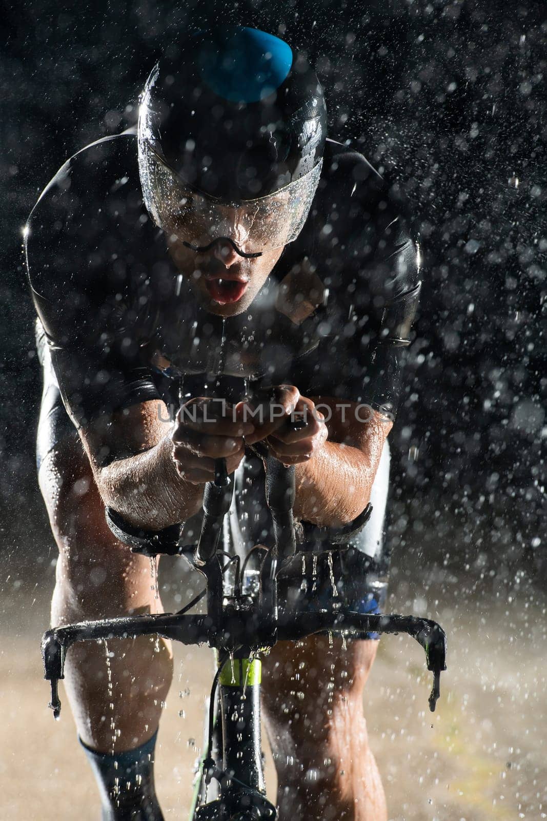 A triathlete braving the rain as he cycles through the night, preparing himself for the upcoming marathon. The blurred raindrops in the foreground and the dark, moody atmosphere in the background add to the sense of determination and grit shown by the athlete. by dotshock