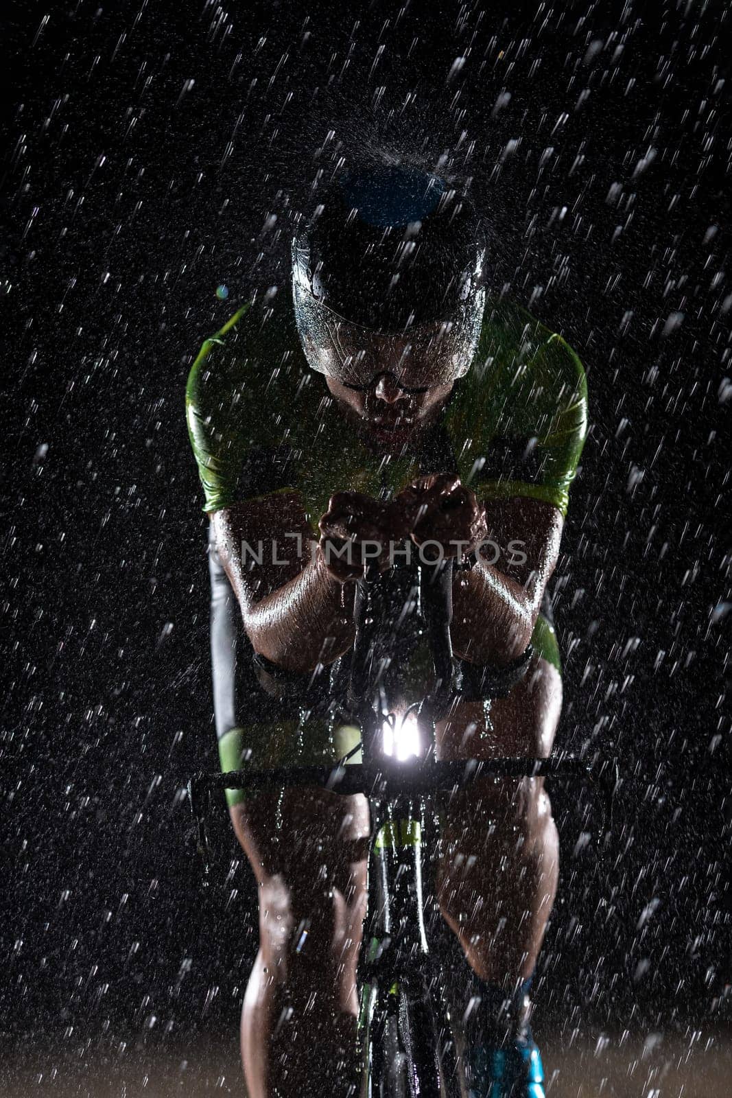 A triathlete braving the rain as he cycles through the night, preparing himself for the upcoming marathon. The blurred raindrops in the foreground and the dark, moody atmosphere in the background add to the sense of determination and grit shown by the athlete