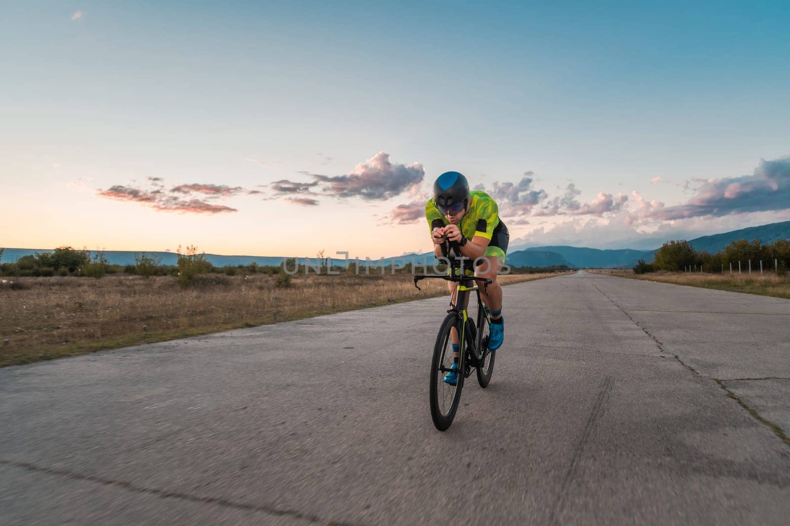 Triathlete riding his bicycle during sunset, preparing for a marathon. The warm colors of the sky provide a beautiful backdrop for his determined and focused effort. by dotshock