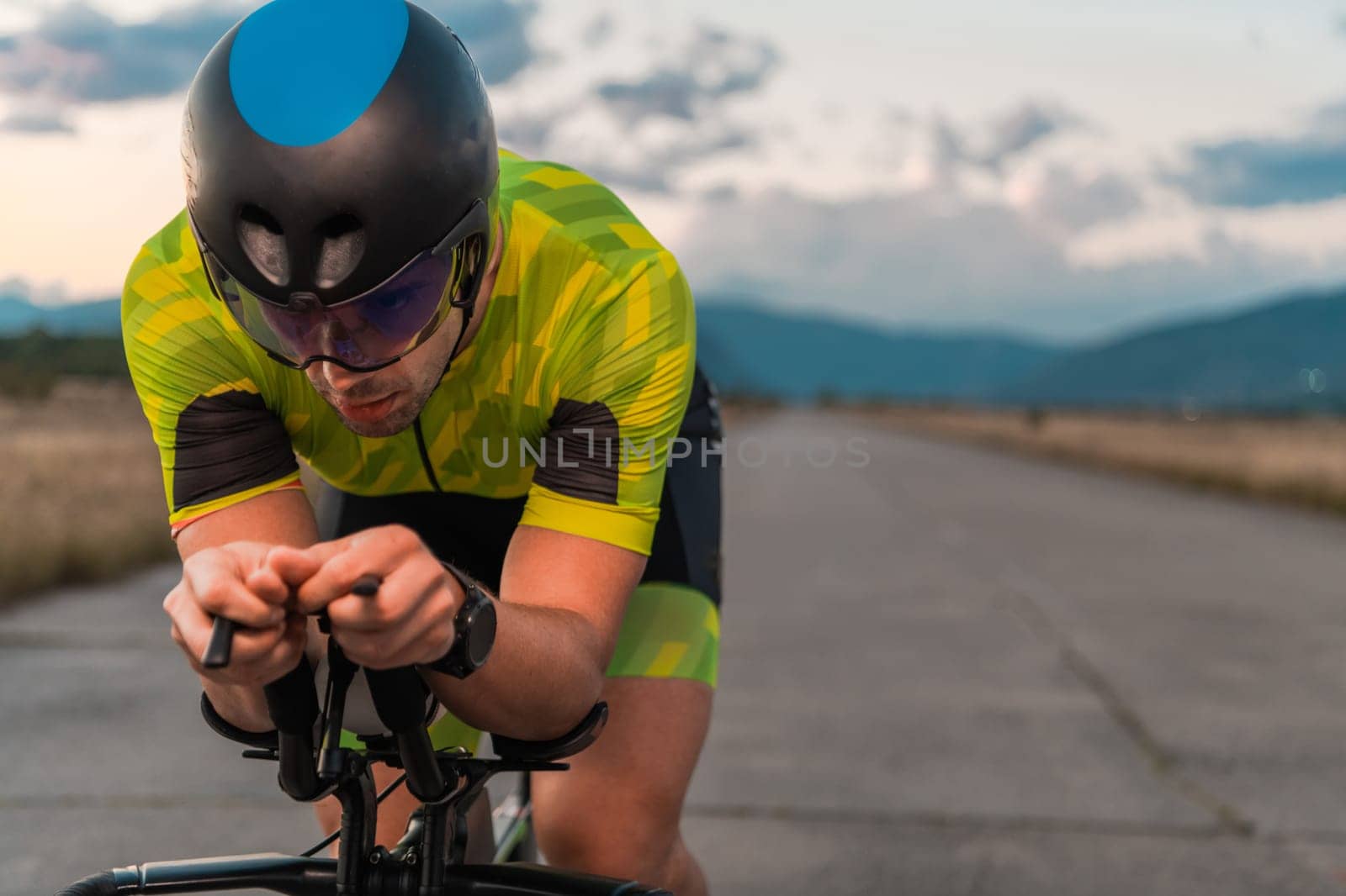 Close up photo of triathlete riding his bicycle during sunset, preparing for a marathon. The warm colors of the sky provide a beautiful backdrop for his determined and focused effort