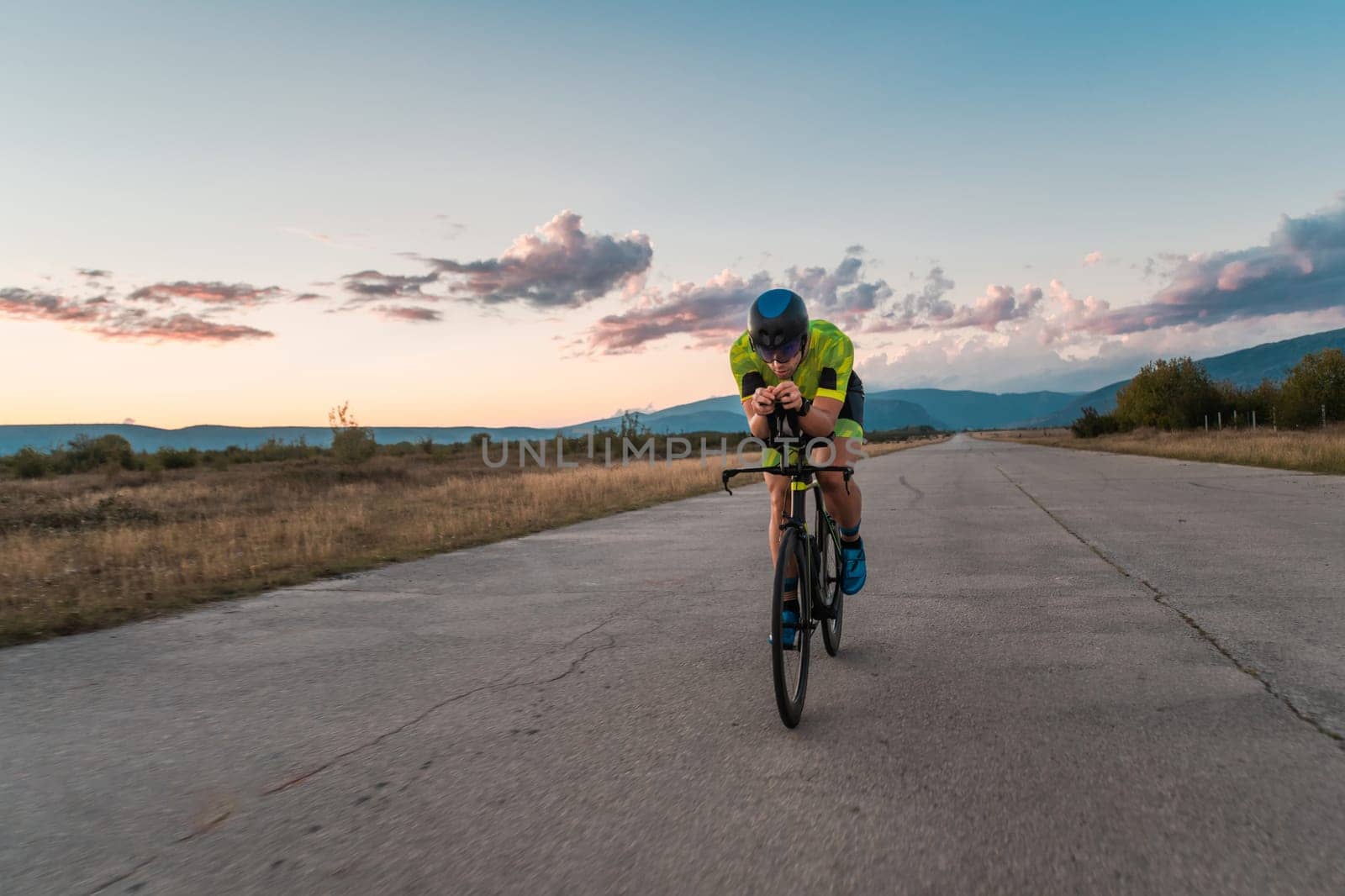 Triathlete riding his bicycle during sunset, preparing for a marathon. The warm colors of the sky provide a beautiful backdrop for his determined and focused effort. by dotshock