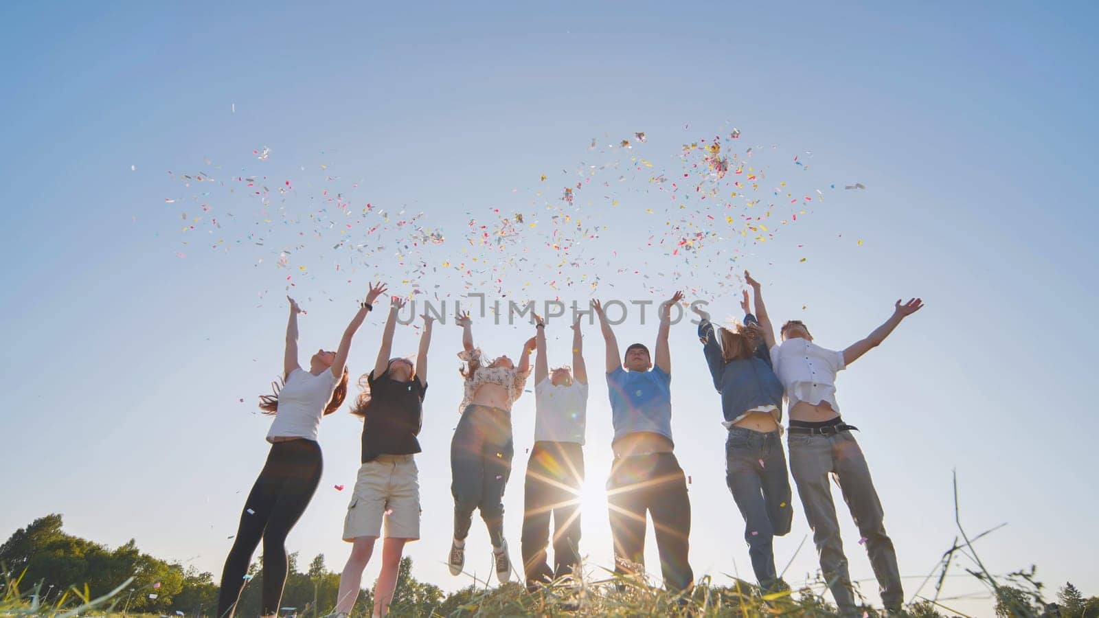 Friends toss colorful paper confetti from their hands against the rays of the evening sun