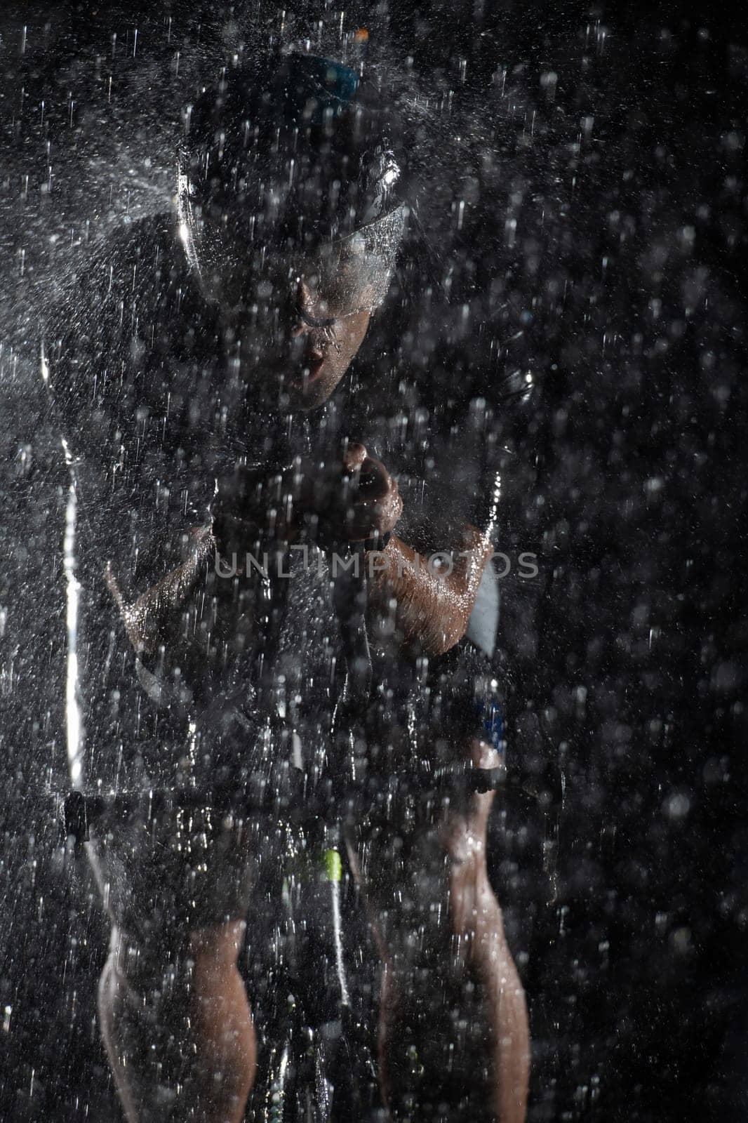 A triathlete braving the rain as he cycles through the night, preparing himself for the upcoming marathon. The blurred raindrops in the foreground and the dark, moody atmosphere in the background add to the sense of determination and grit shown by the athlete