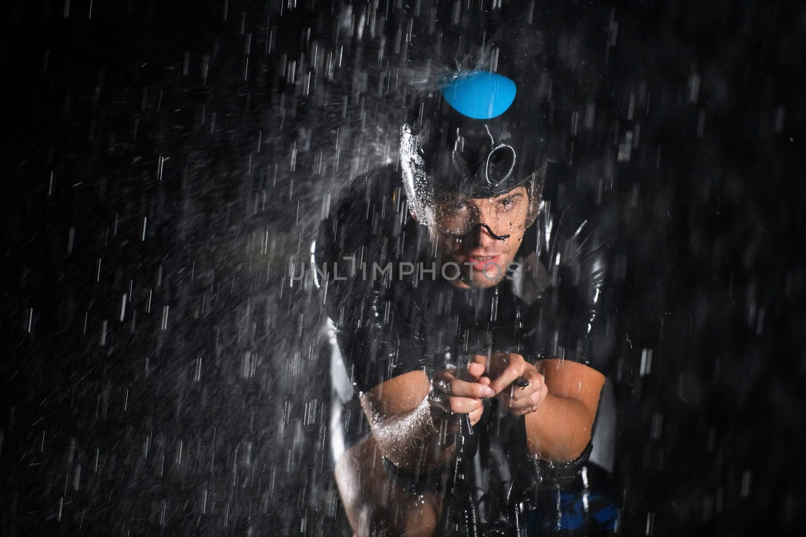 A triathlete braving the rain as he cycles through the night, preparing himself for the upcoming marathon. The blurred raindrops in the foreground and the dark, moody atmosphere in the background add to the sense of determination and grit shown by the athlete