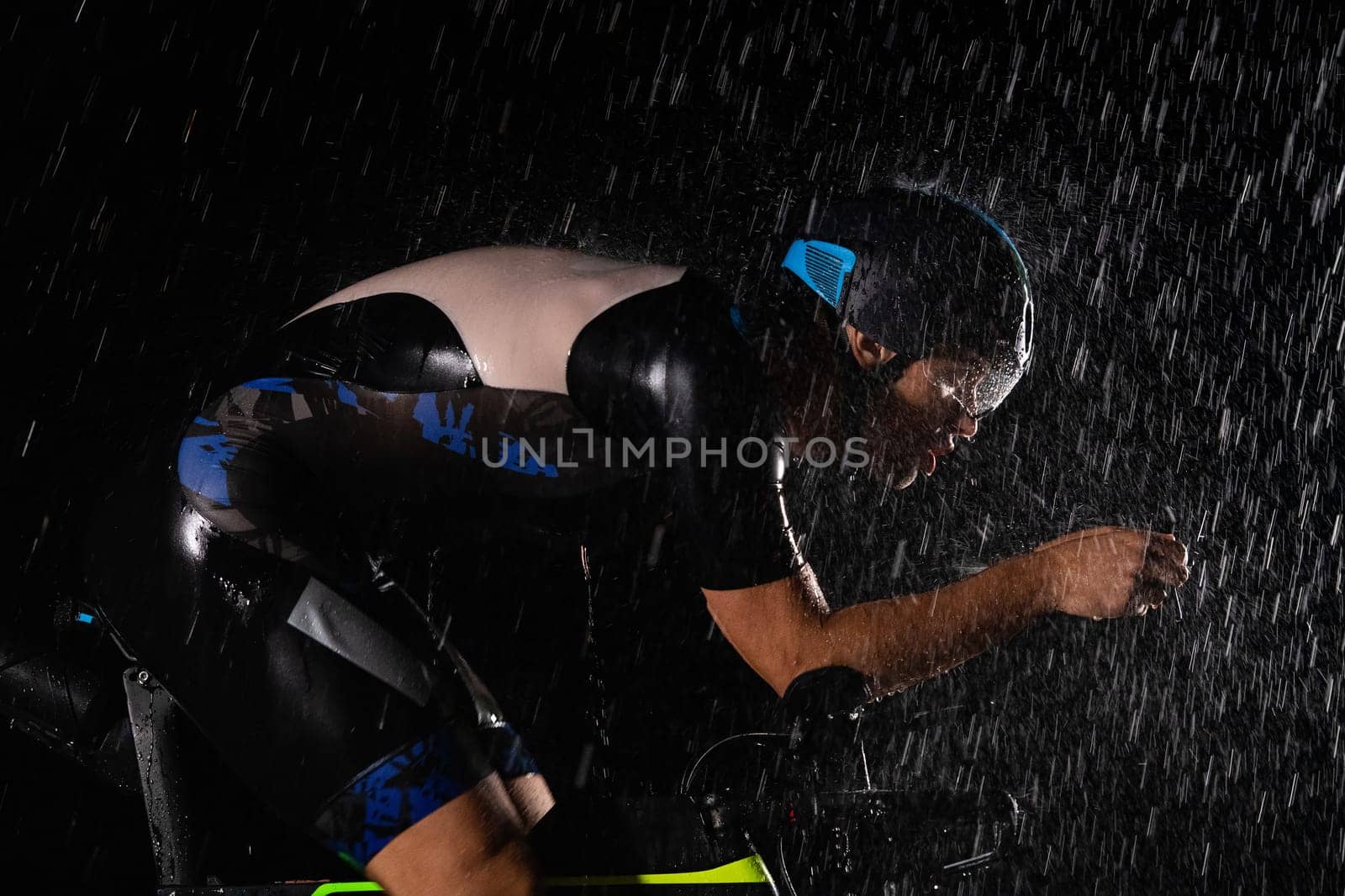 A triathlete braving the rain as he cycles through the night, preparing himself for the upcoming marathon. The blurred raindrops in the foreground and the dark, moody atmosphere in the background add to the sense of determination and grit shown by the athlete. by dotshock