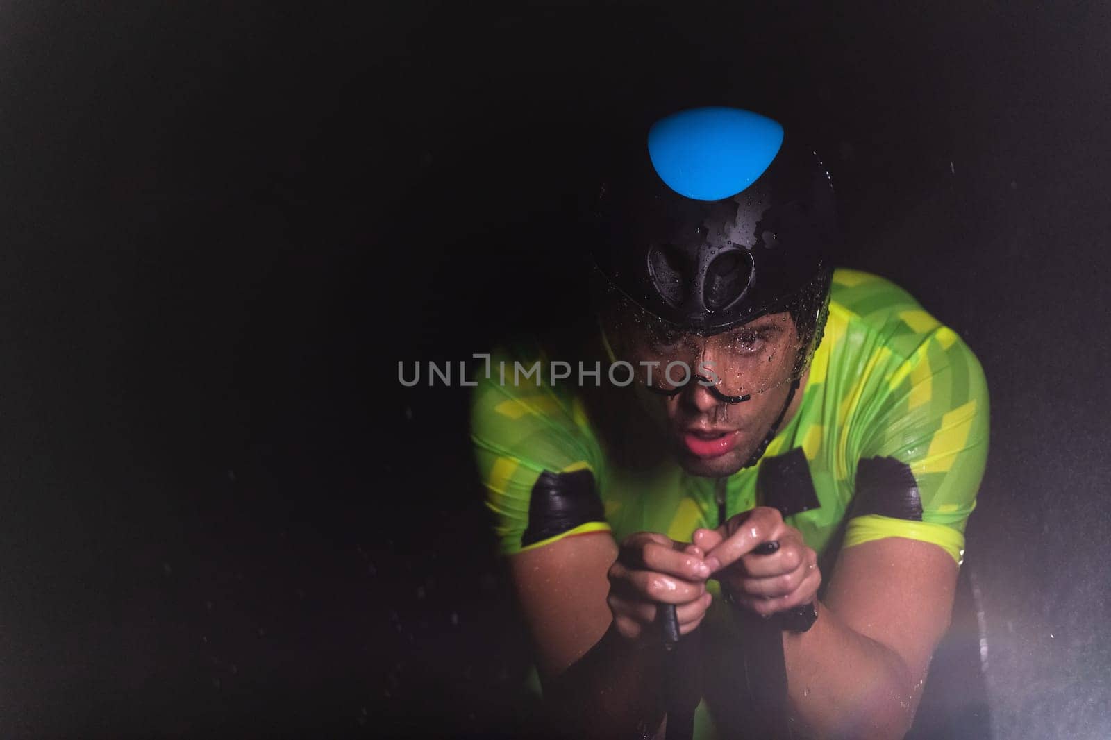 A triathlete braving the rain as he cycles through the night, preparing himself for the upcoming marathon. The blurred raindrops in the foreground and the dark, moody atmosphere in the background add to the sense of determination and grit shown by the athlete. by dotshock