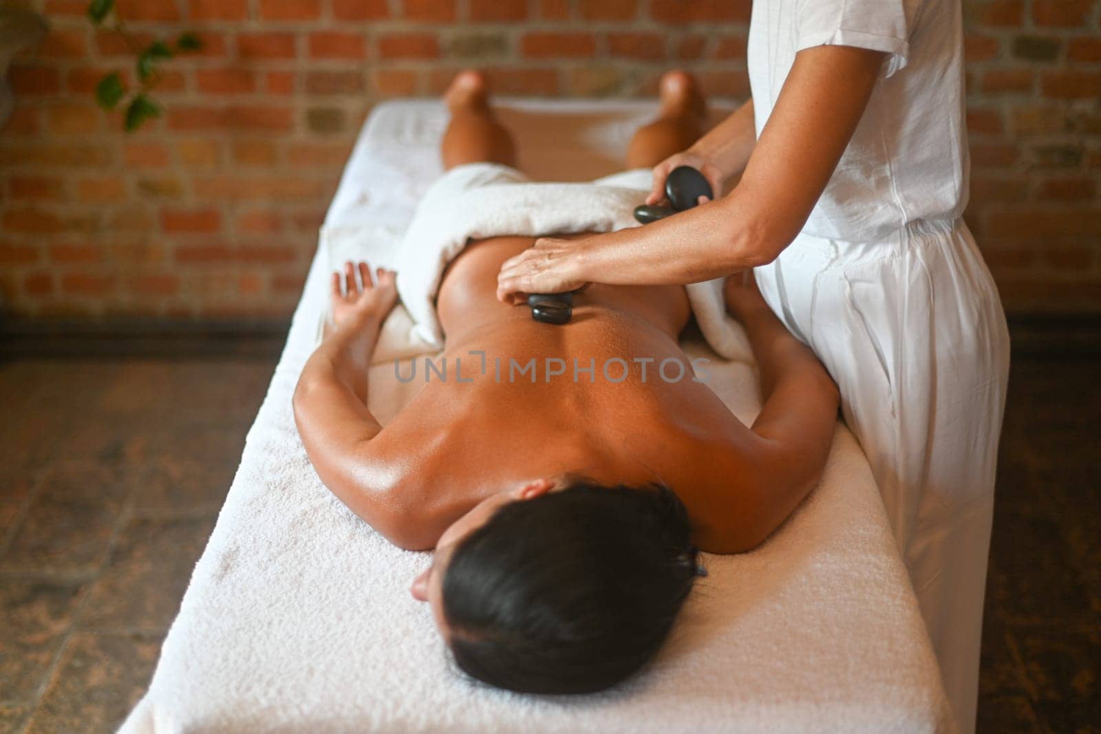 professional caucasian masseuse physiotherapist performing stone massage to hispanic brunette woman in spa clinic by verbano