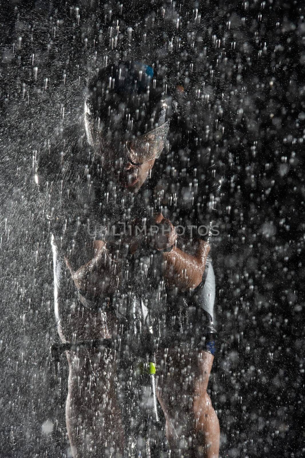 A triathlete braving the rain as he cycles through the night, preparing himself for the upcoming marathon. The blurred raindrops in the foreground and the dark, moody atmosphere in the background add to the sense of determination and grit shown by the athlete