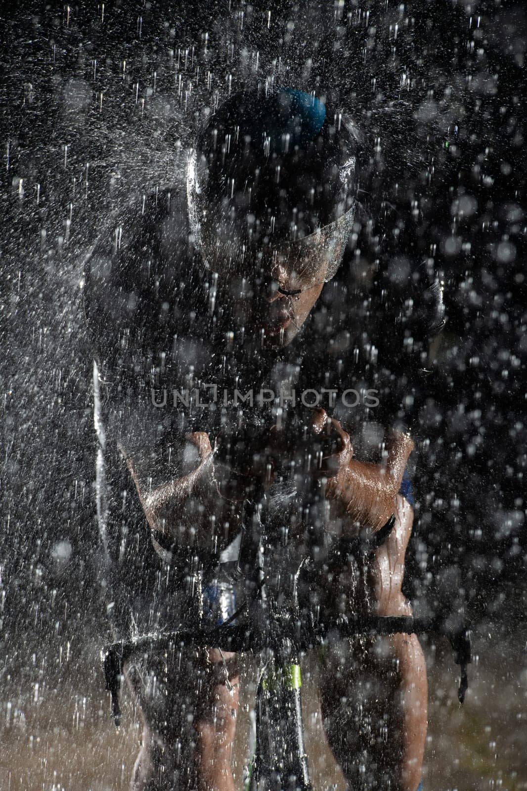 A triathlete braving the rain as he cycles through the night, preparing himself for the upcoming marathon. The blurred raindrops in the foreground and the dark, moody atmosphere in the background add to the sense of determination and grit shown by the athlete