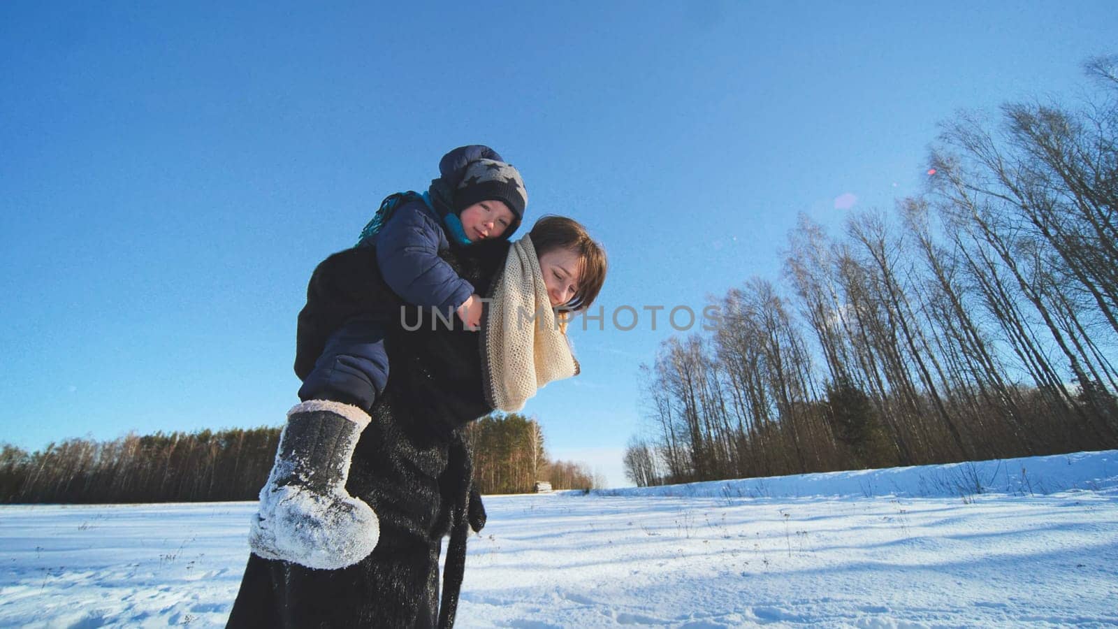 A mother carries her son across a field on her shoulders in winter