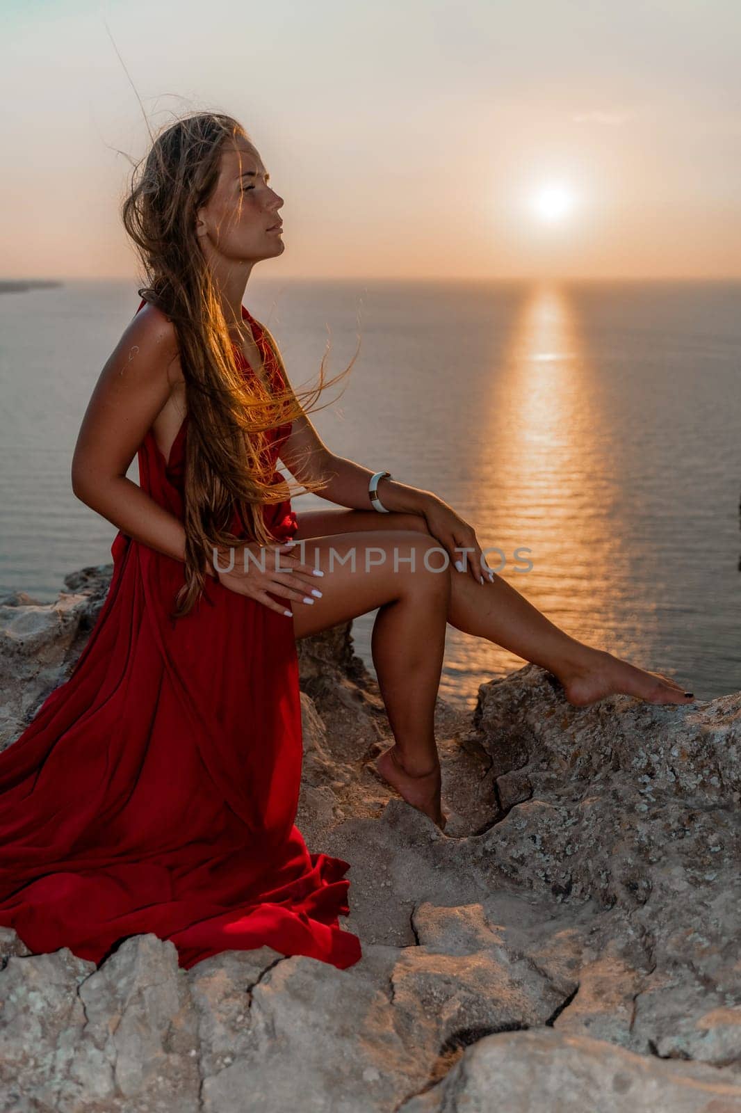 Woman sunset sea red dress, side view a happy beautiful sensual woman in a red long dress posing on a rock high above the sea on sunset