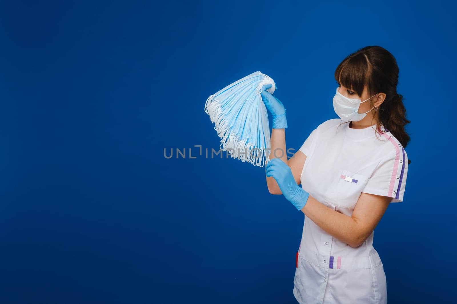 A young girl doctor in a medical mask holds a lot of masks in her hands. A nurse with white masks in her hands on a blue background by Lobachad