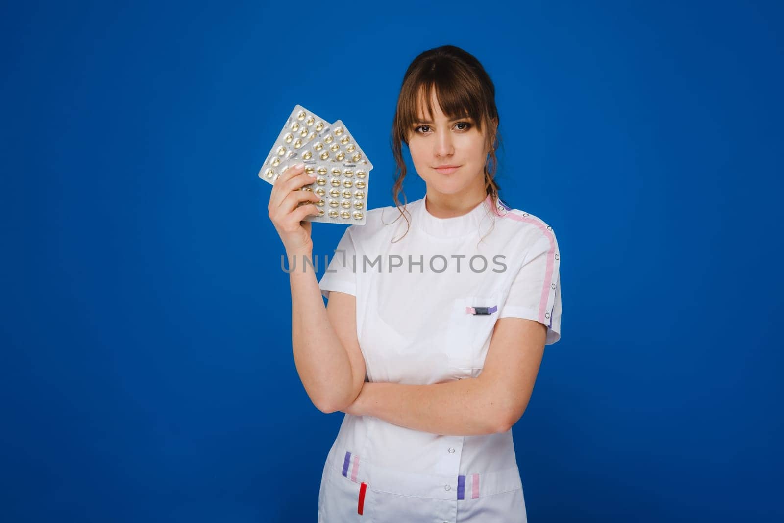 The concept of health care. A young brunette doctor in a white coat on a blue background shows plates with capsules to take. by Lobachad