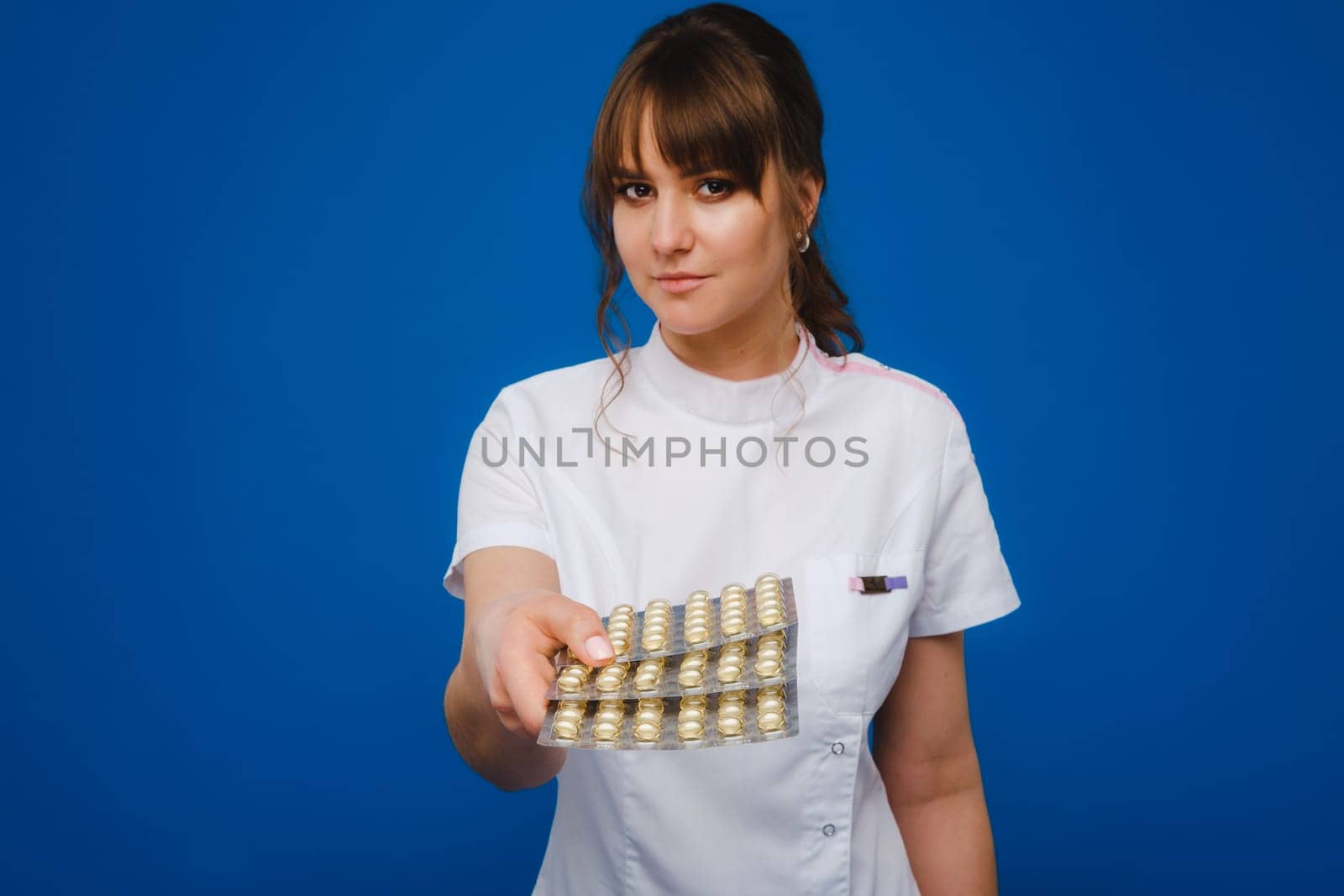 The concept of health care. A young brunette doctor in a white coat on a blue background shows plates with capsules to take. by Lobachad
