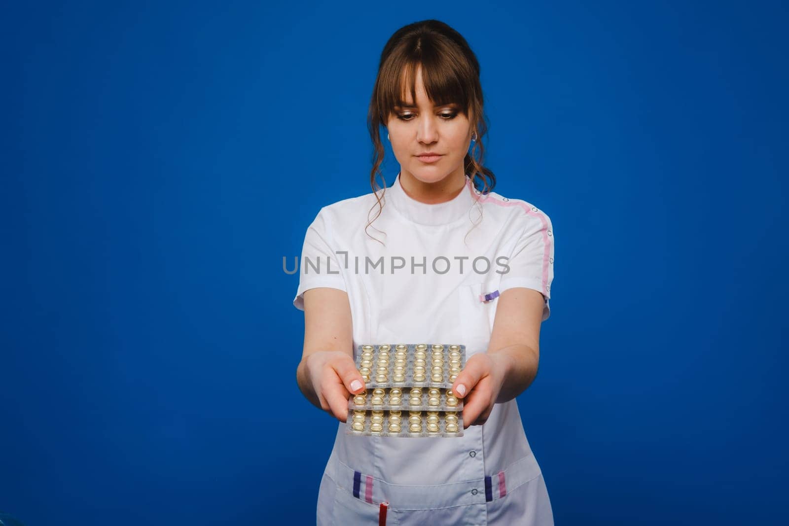 The concept of health care. A young brunette doctor in a white coat on a blue background shows plates with capsules to take. by Lobachad