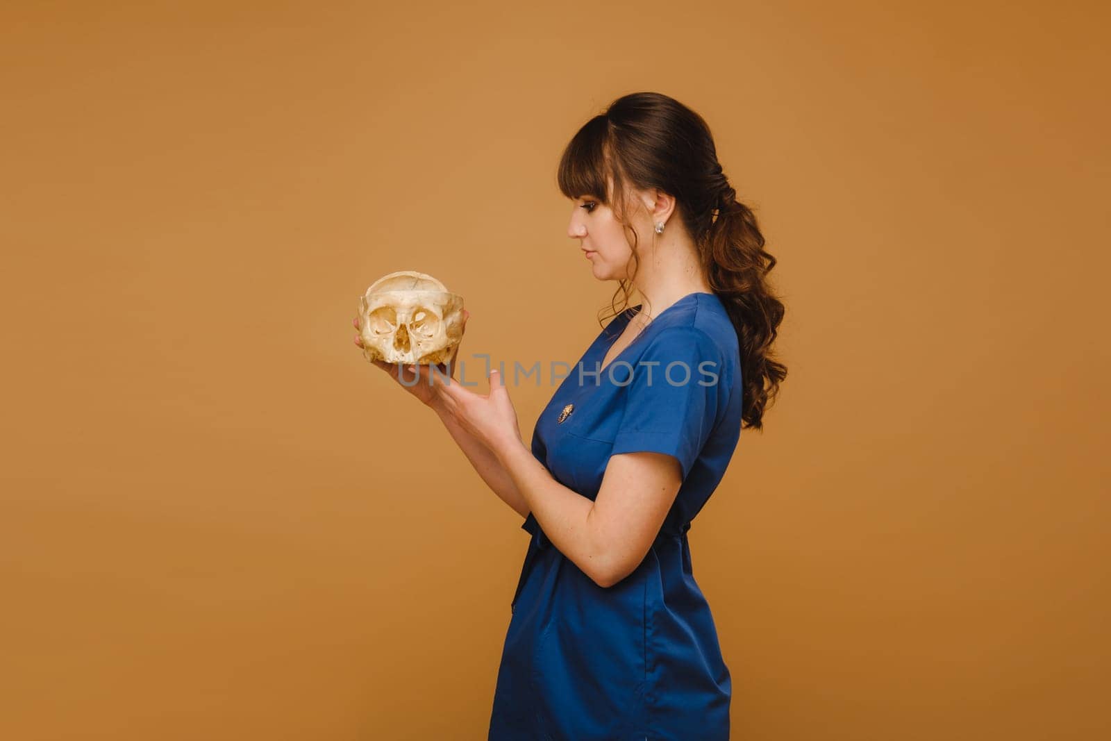 Cute young doctor girl holding a human skull, brown background behind the doctor.