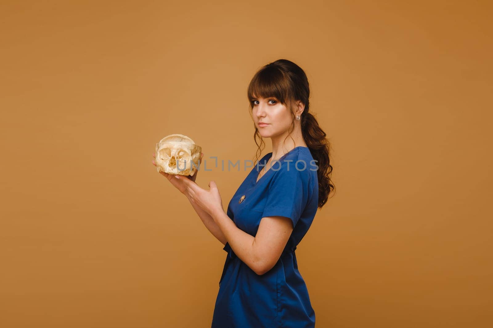 Cute young doctor girl holding a human skull, brown background behind the doctor.