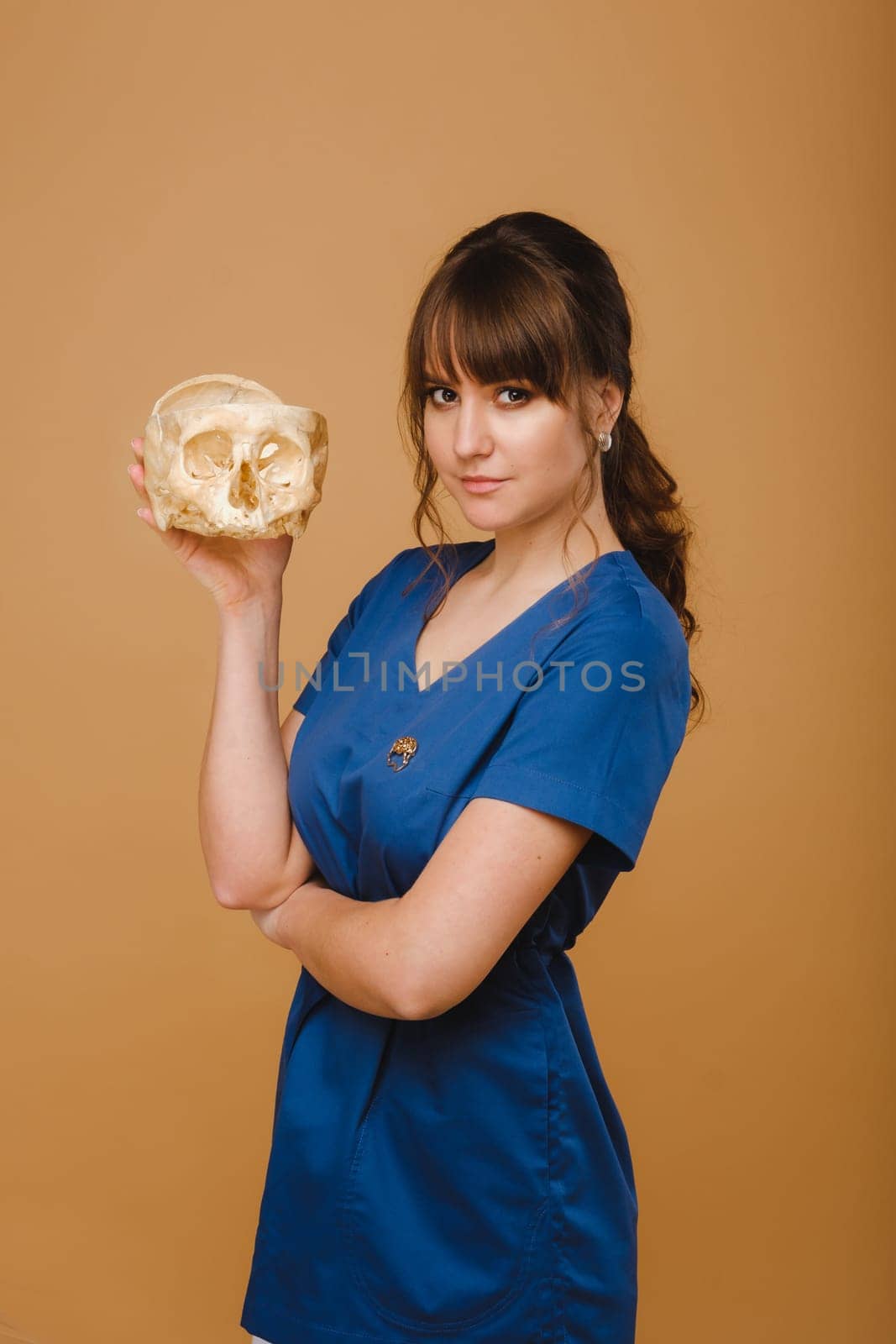 Cute young doctor girl holding a human skull, brown background behind the doctor.