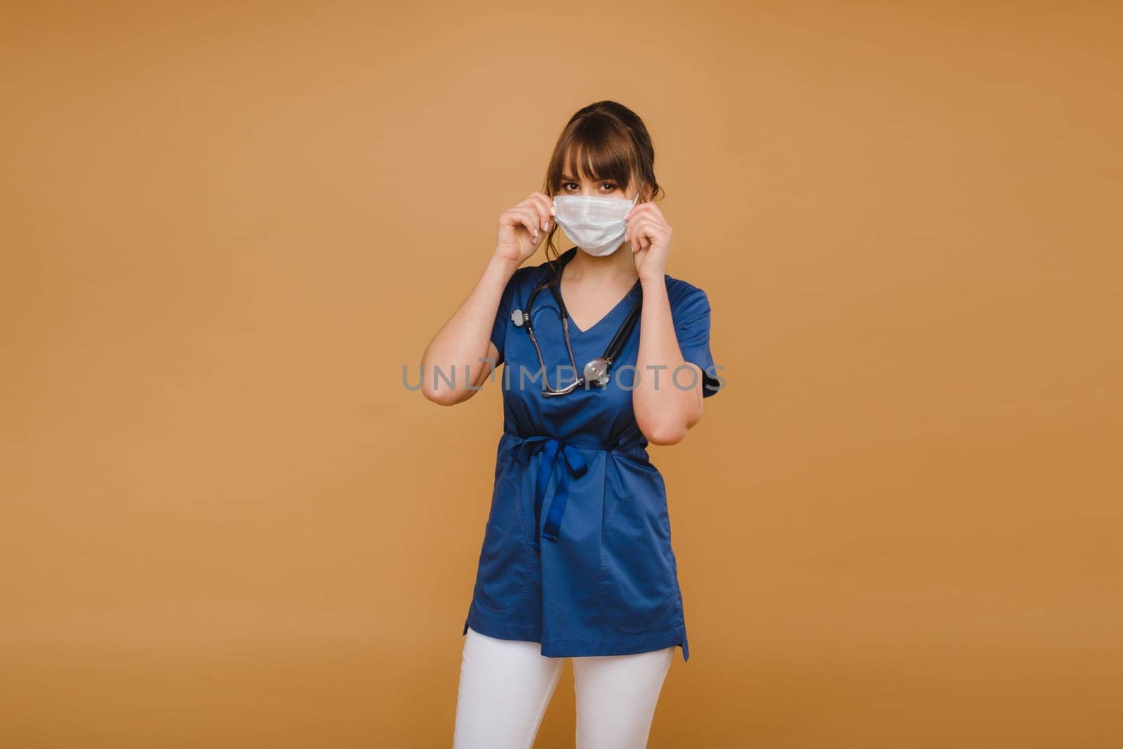 A girl doctor stands in a medical mask, isolated on a brown background by Lobachad
