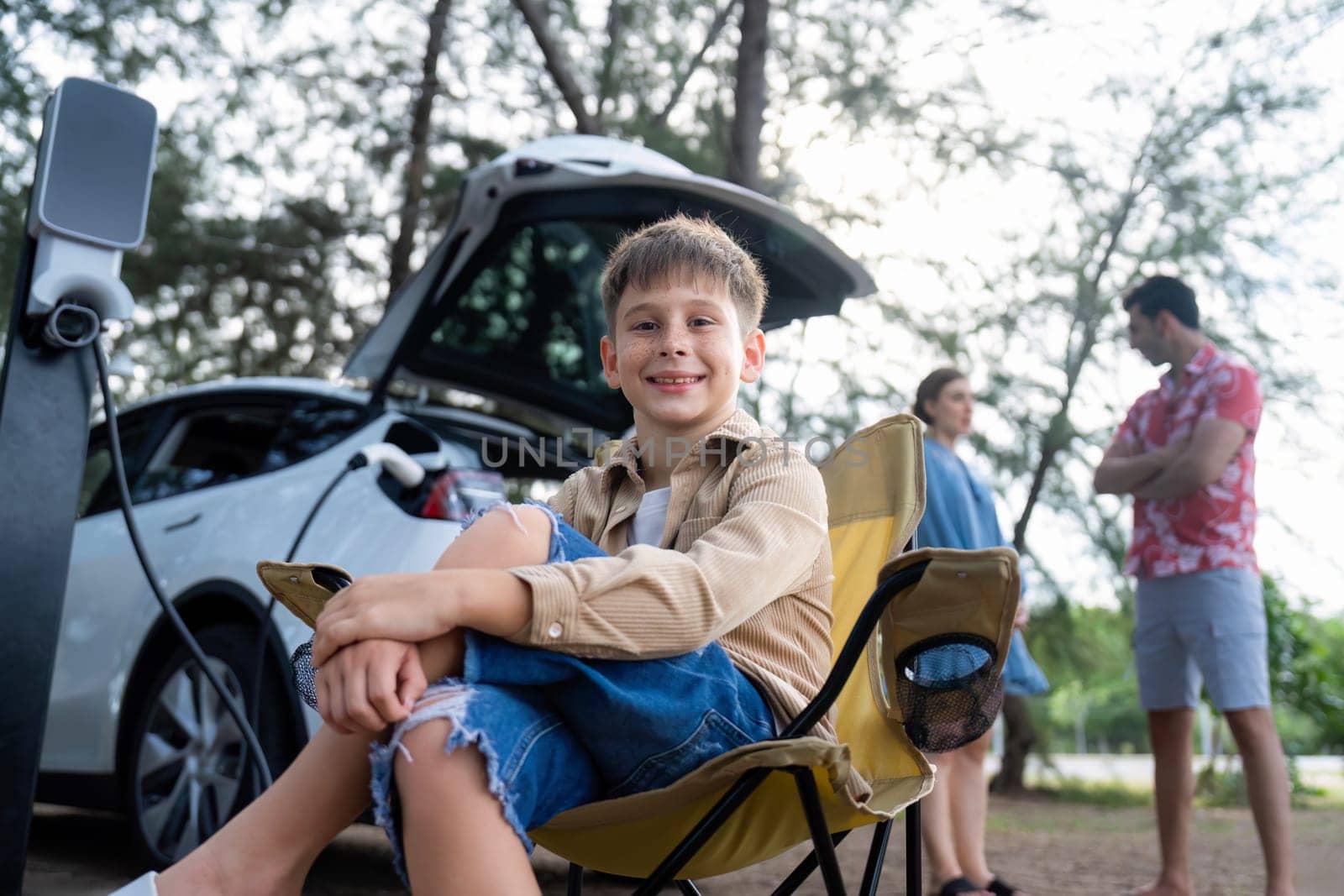 Little boy portrait sitting on camping chair with his family in background. Road trip travel with alternative energy charging station for eco-friendly car concept. Perpetual
