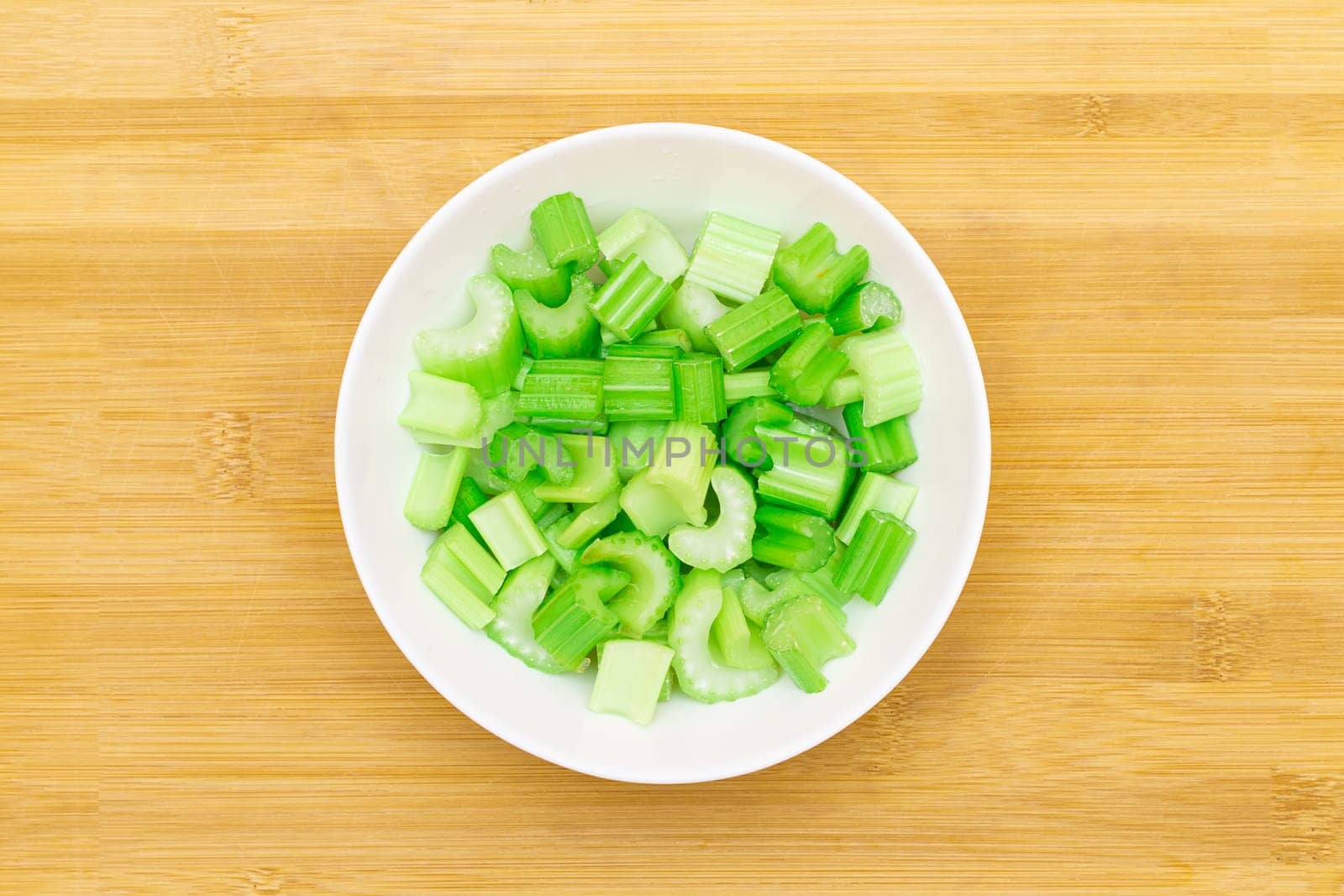 Fresh Chopped Celery Slices in White Bowl on Bamboo Cutting Board - Top View. Vegan and Vegetarian Culture. Raw Food. Healthy Diet with Negative Calorie Content