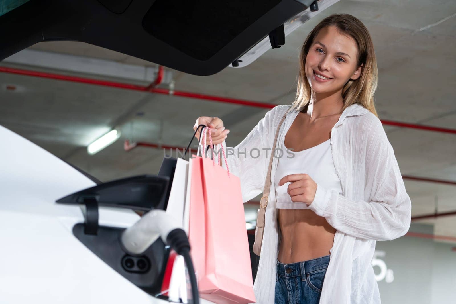 Young woman travel with EV electric car to shopping center parking lot charging in downtown city showing urban sustainability lifestyle by green clean rechargeable energy of electric vehicle innards