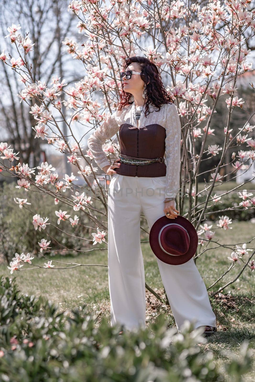 Magnolia park woman. Stylish woman in a hat stands near the magnolia bush in the park. Dressed in white corset pants and posing for the camera