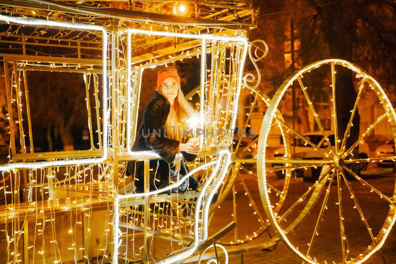 Woman holding sparkler night while celebrating Christmas outside. Dressed in a fur coat and a red headband. Blurred christmas decorations in the background. Selective focus.