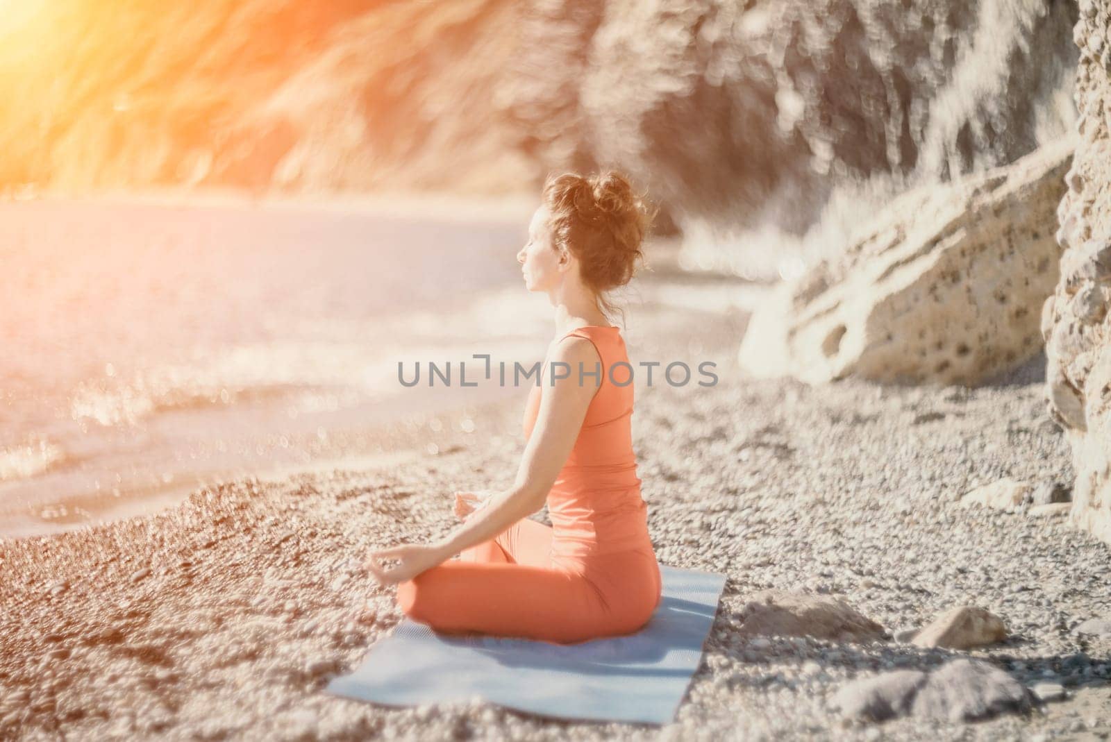 The woman in a red suit practicing yoga on stone at sunrise near the sea. Young beautiful girl in a red bathing suit sits on the seashore in lotus position. Yoga. Healthy lifestyle. Meditation