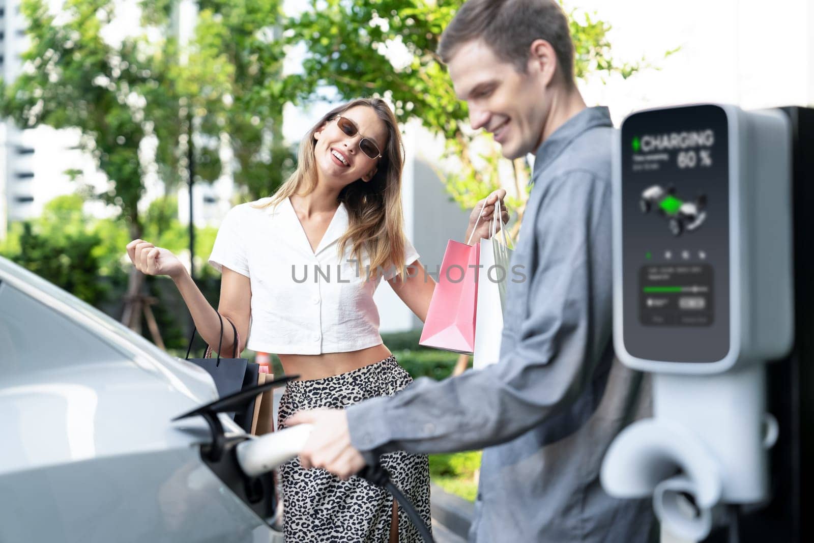 Young couple travel with EV electric car charging in green sustainable city outdoor garden in summer shows urban sustainability lifestyle by green clean rechargeable energy of electric vehicle innards