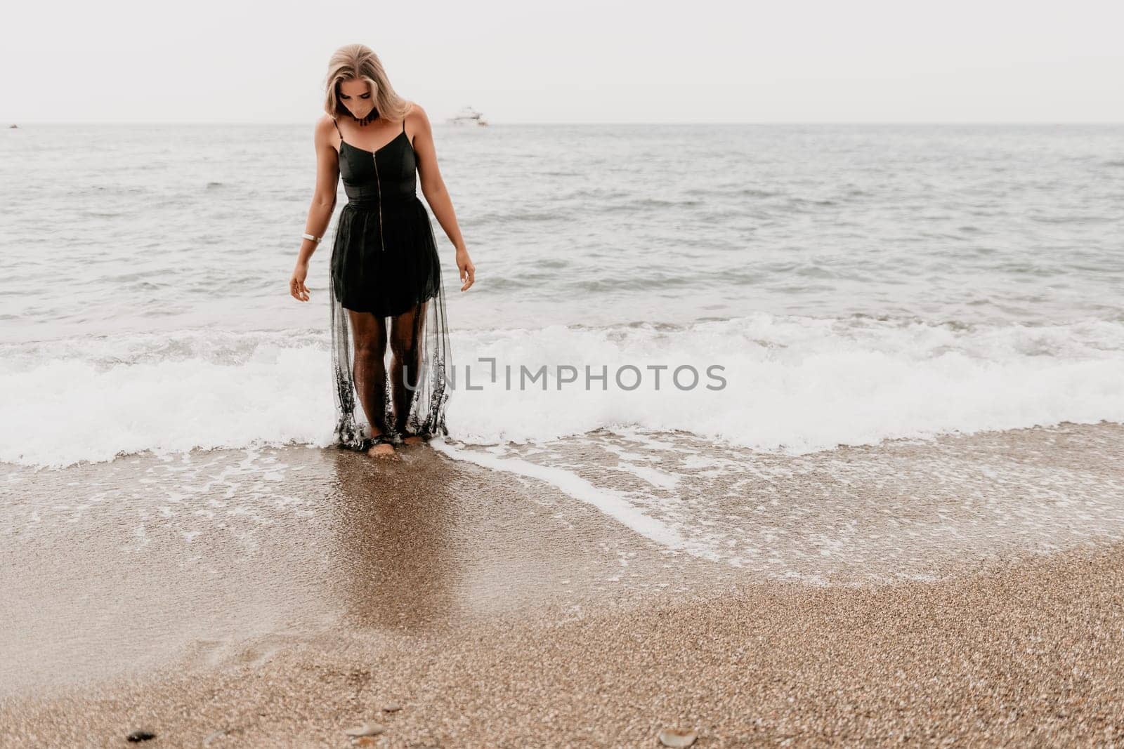 Woman summer travel sea. Happy tourist in black dress enjoy taking picture outdoors for memories. Woman traveler posing on sea beach surrounded by volcanic mountains, sharing travel adventure journey by panophotograph