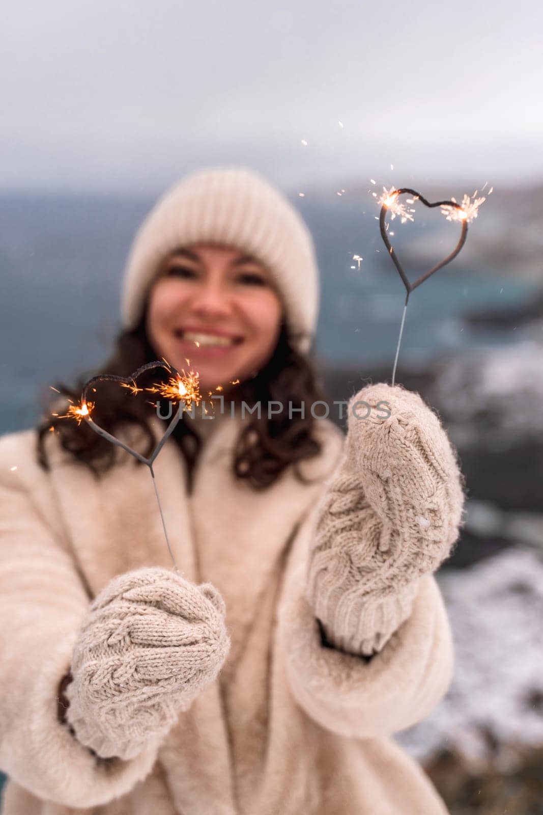 Outdoor winter portrait of happy smiling woman, light faux fur coat holding heart sparkler, posing against sea and snow background by Matiunina