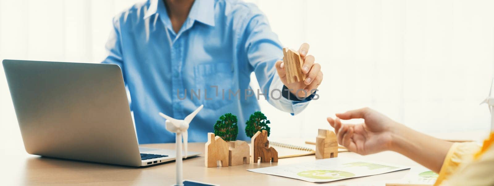 Two business people talking about eco city at green business meeting on meeting table with windmill represented renewable energy, wooden house block and tree model represent green city. Delineation.