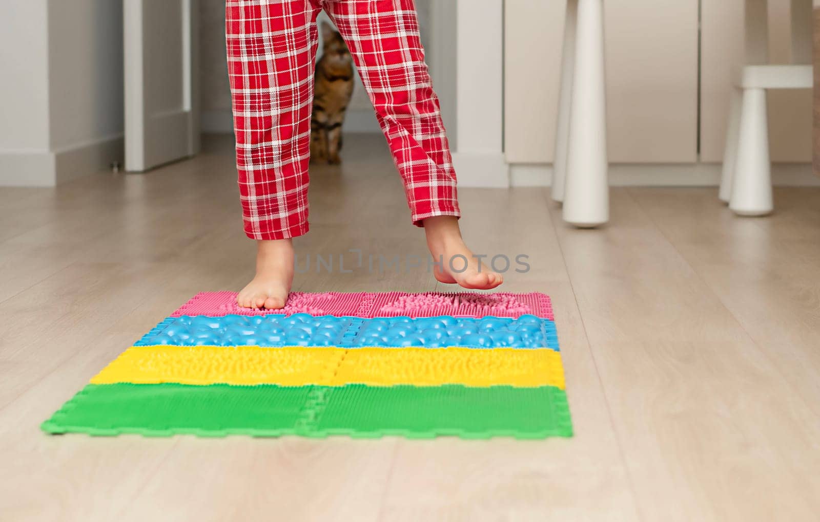 Sport and health concept. A little boy 4 years old in red checkered pajamas is working out on a multi-colored massage orthopedic mat with spikes in a home interior. Close-up. by ketlit