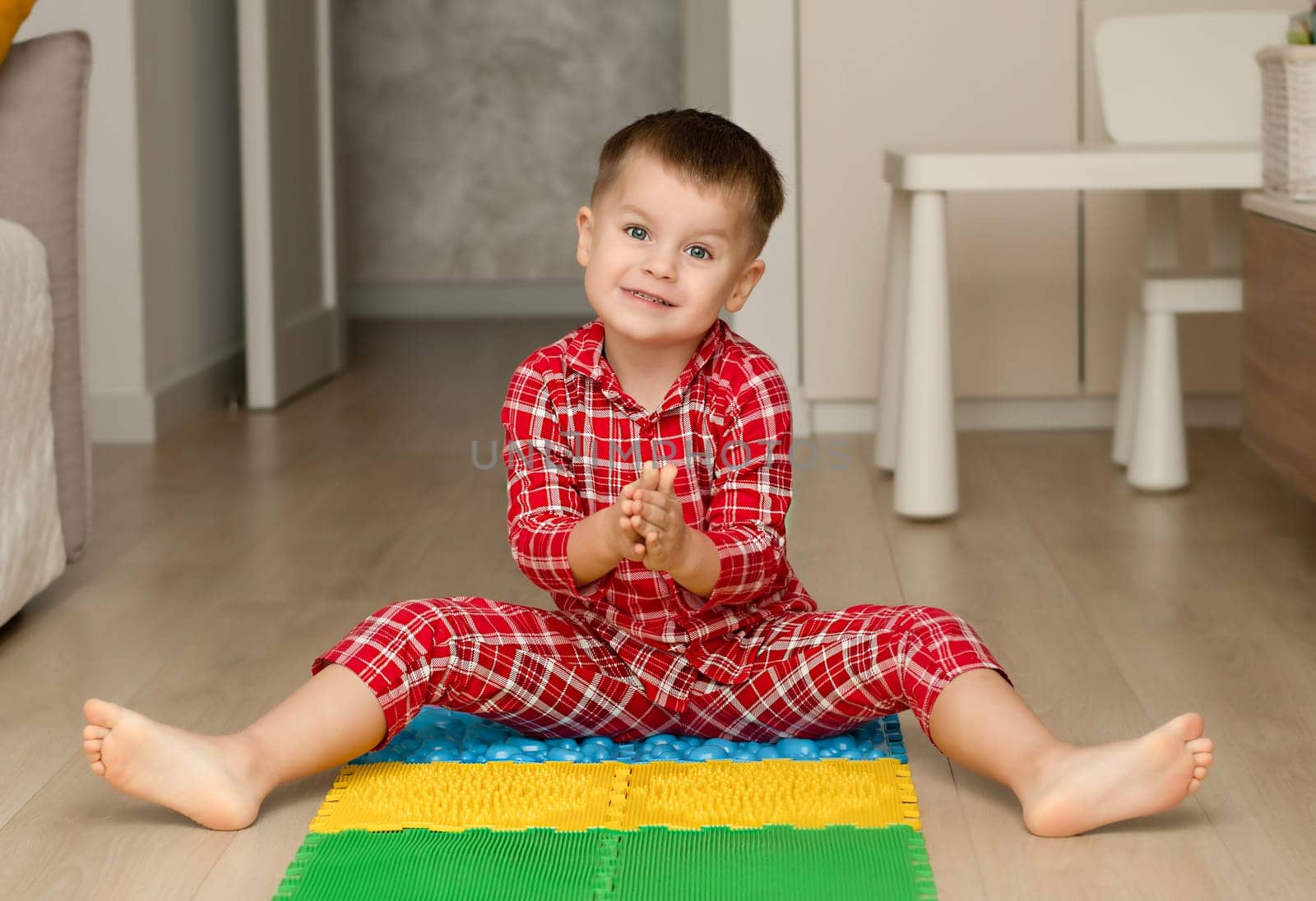 Sport and health concept. A little boy 4 years old in red checkered pajamas is having fun on a multi-colored massage orthopedic mat with spikes in a home interior. Close-up. Soft focus.