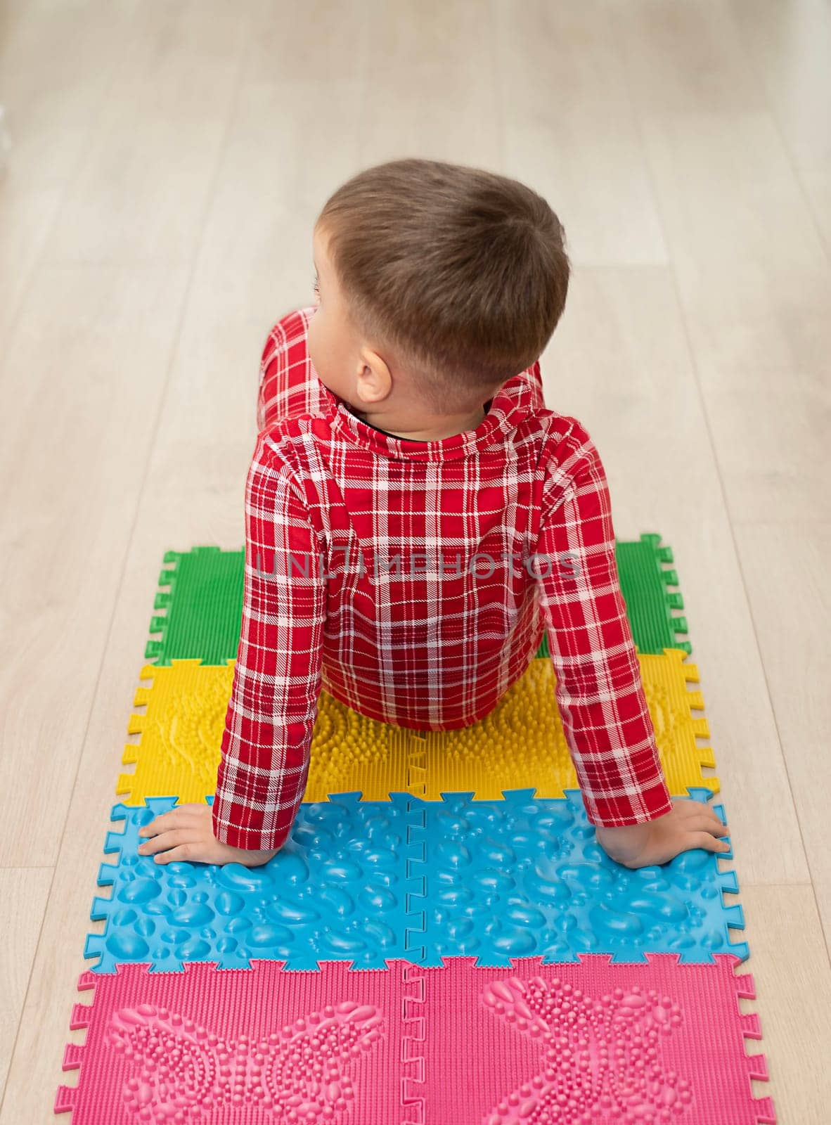 Sport and health concept. A little boy 4 years old in red checkered pajamas is having fun on a multi-colored massage orthopedic mat with spikes in a home interior. Close-up. Soft focus.