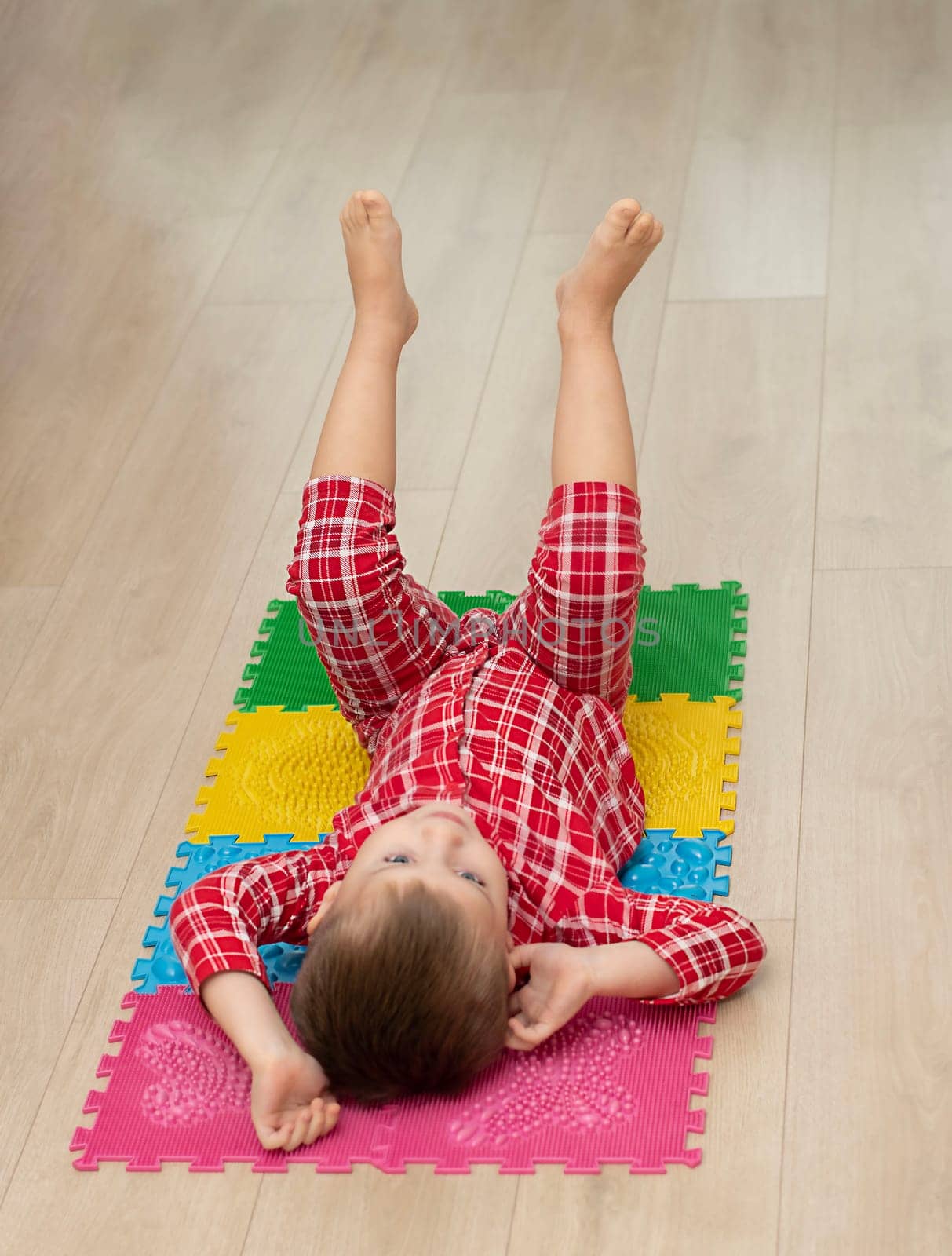 Sport and health concept. A little boy 4 years old in red checkered pajamas is working out on a multi-colored massage orthopedic mat with spikes in a home interior. Close-up. by ketlit