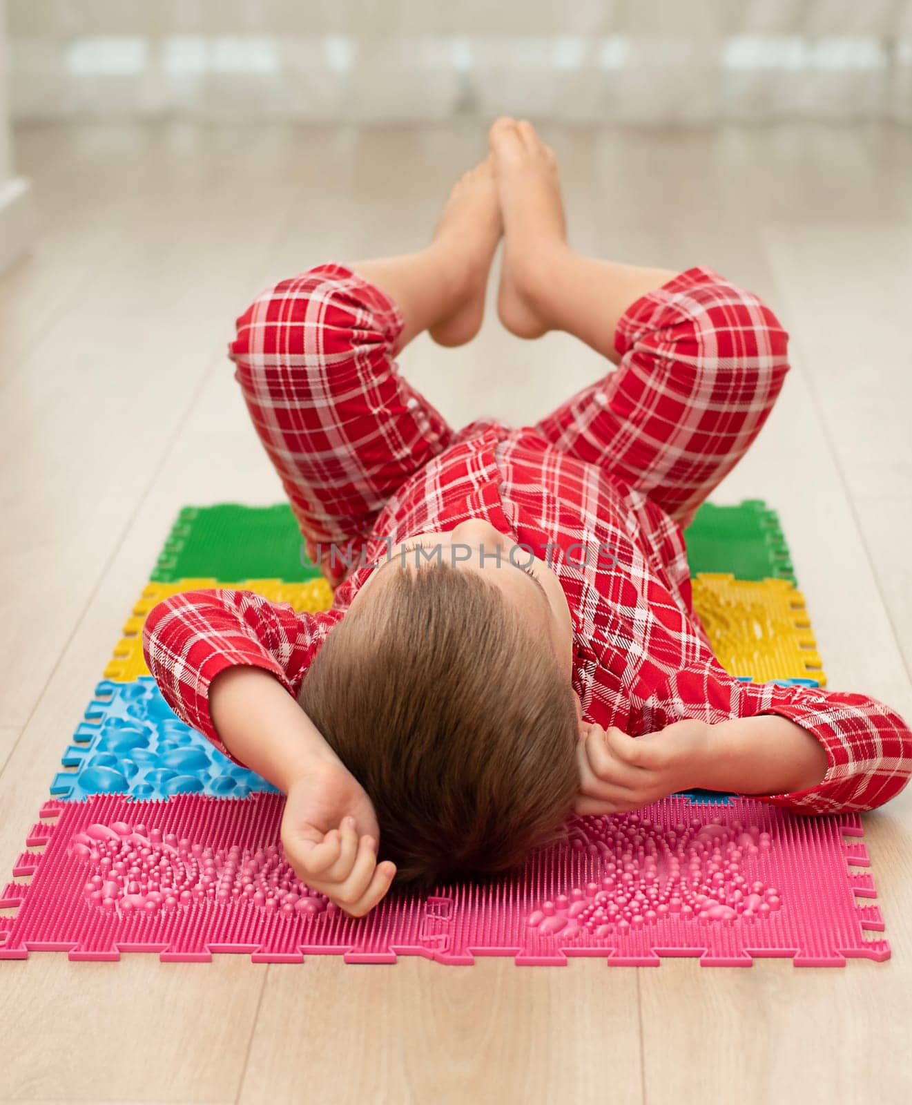 Sport and health concept. A little boy 4 years old in red checkered pajamas is working out on a multi-colored massage orthopedic mat with spikes in a home interior. Close-up. by ketlit