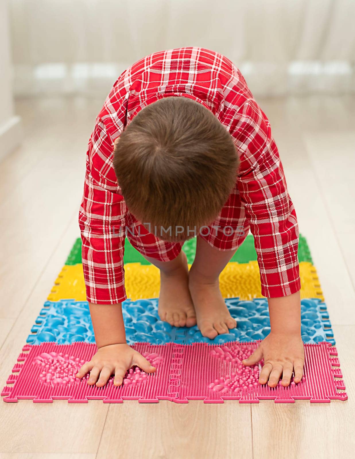 Sport and health concept. A little boy 4 years old in red checkered pajamas is working out on a multi-colored massage orthopedic mat with spikes in a home interior. Close-up. by ketlit