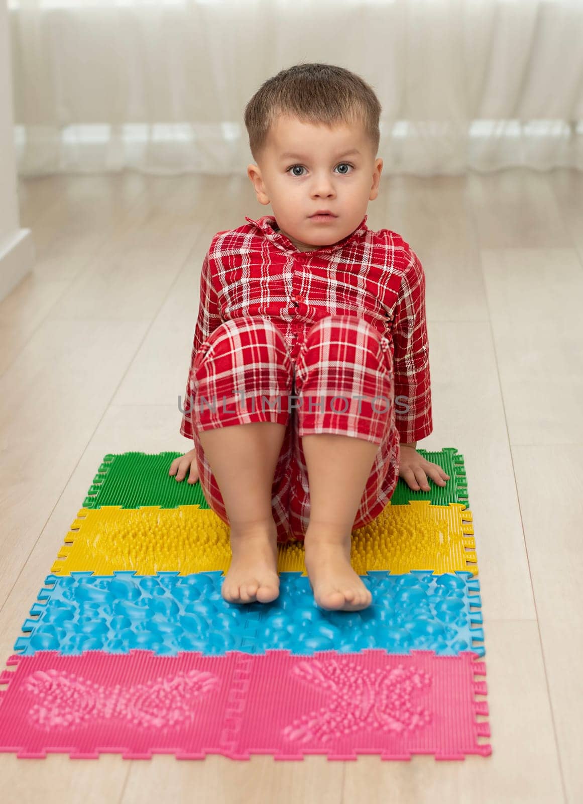 Sport and health concept. A little boy 4 years old in red checkered pajamas is having fun on a multi-colored massage orthopedic mat with spikes in a home interior. Close-up. Soft focus.