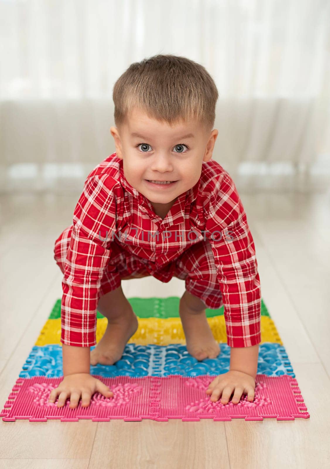 Sport and health concept. A little boy 4 years old in red checkered pajamas is having fun on a multi-colored massage orthopedic mat with spikes in a home interior. Close-up. Soft focus.