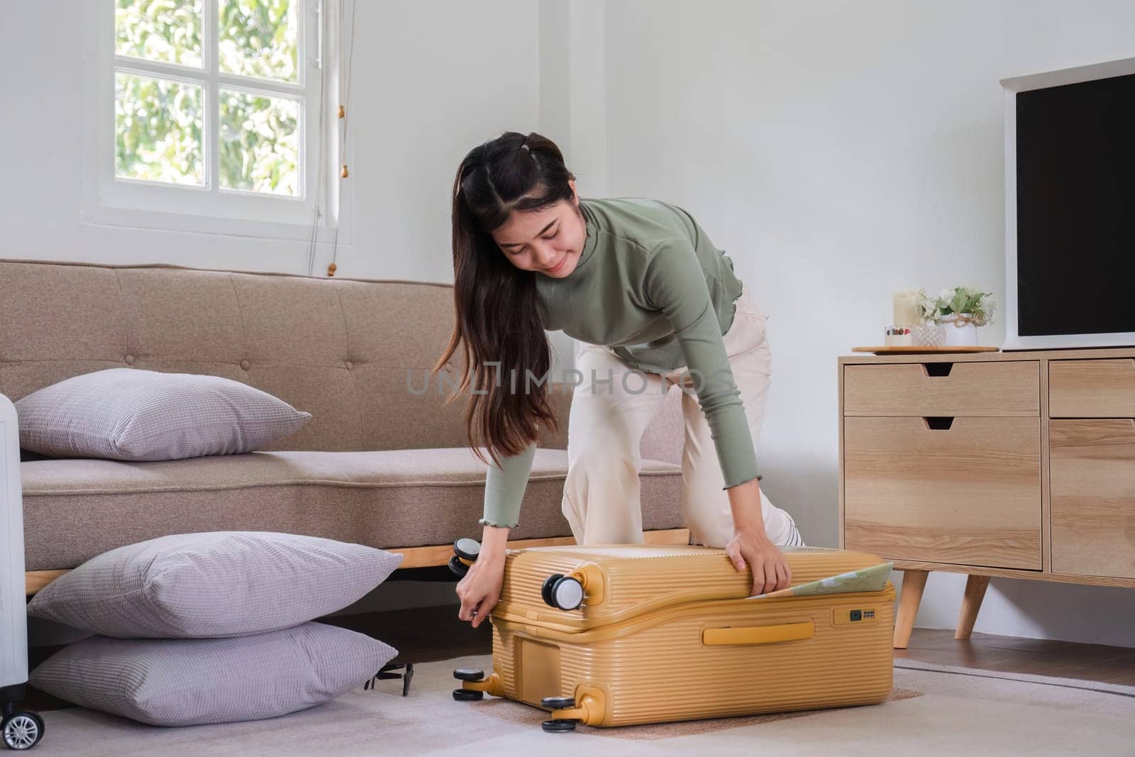 Beautiful Asian woman try to pack clothes into a yellow suitcase to prepare for a weekend getaway. by wichayada