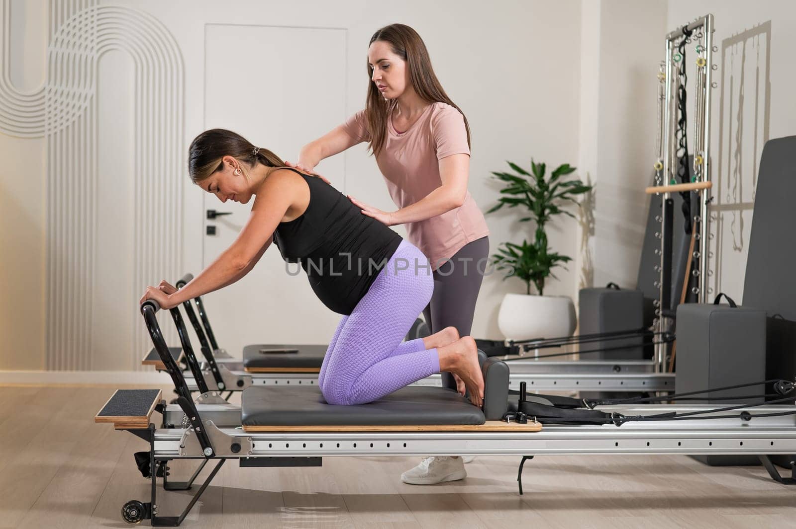 A pregnant woman works out on a reformer exercise machine with a personal trainer